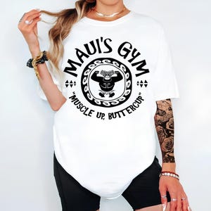 May include: White t-shirt with a black graphic of a muscular figure inside a circular design. The text "MAUI'S GYM" arches above the figure, with "MUSCLE UP, BUTTERCUP" below. The shirt is a casual, oversized fit.
