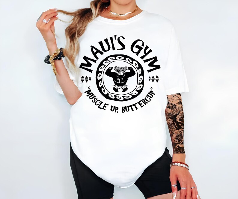 May include: White t-shirt with a black graphic of a muscular figure inside a circular design. The text "MAUI'S GYM" arches above the figure, with "MUSCLE UP, BUTTERCUP" below. The shirt is a casual, oversized fit.