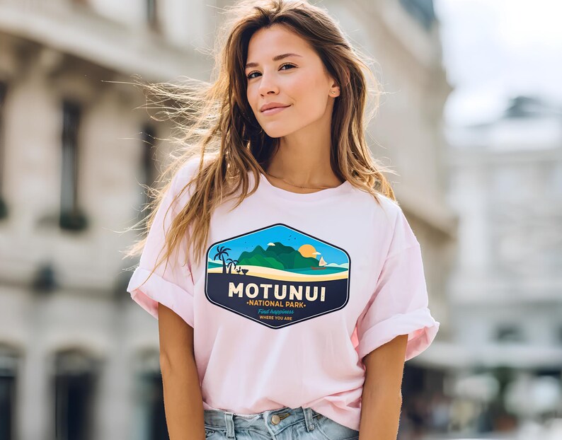 May include: A pale pink t-shirt with rolled sleeves. The shirt displays a colourful graphic of a landscape with the text "MOTUNUI - NATIONAL PARK - Where You Are". The graphic includes a beach, mountains, and a setting sun.