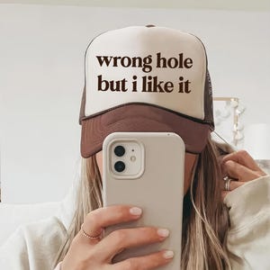 May include: A brown and white trucker hat with the text "wrong hole but I like it" in black. The hat has a mesh back and a curved brim. The hat is being worn by a person.
