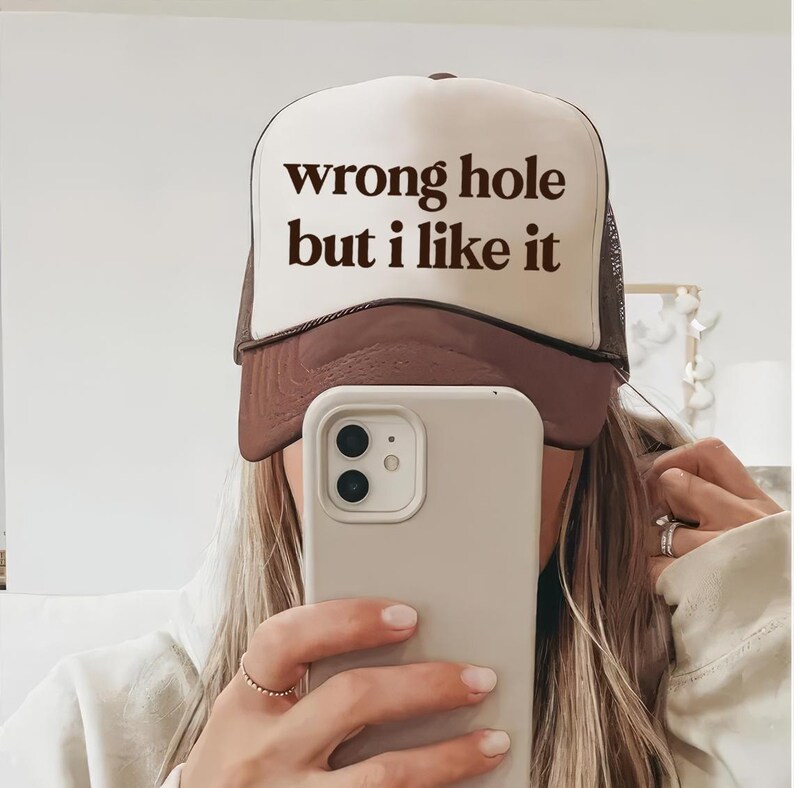 May include: A brown and white trucker hat with the text "wrong hole but I like it" in black. The hat has a mesh back and a curved brim. The hat is being worn by a person.