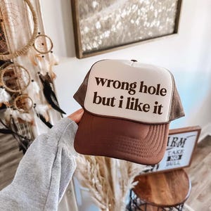 May include: A brown trucker hat with a white front panel. The text "wrong hole but i like it" is printed in dark brown on the front. The hat has a brown mesh back and a curved brim.