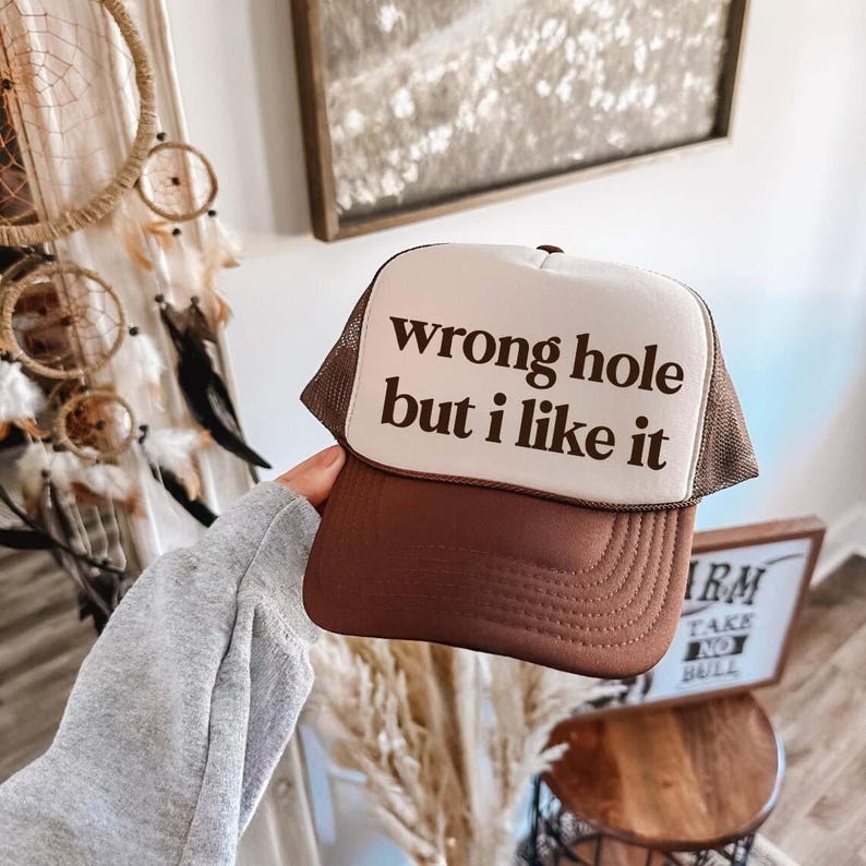 May include: A brown trucker hat with a white front panel. The text "wrong hole but i like it" is printed in dark brown on the front. The hat has a brown mesh back and a curved brim.