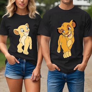 May include: Black t-shirts featuring Simba and Nala from The Lion King. The Simba shirt is worn by a man, and the Nala shirt is worn by a woman. Both shirts have a crew neck and short sleeves. The cartoon lion cubs are in shades of yellow and brown.