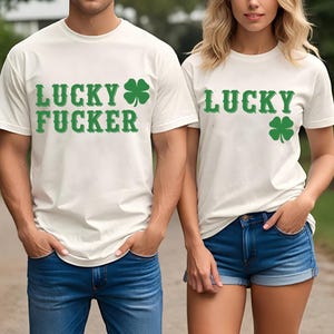 May include: Off-white t-shirts with green text and shamrock designs. One shirt reads "LUCKY FUCKER" and the other reads "LUCKY". Both shirts are paired with blue denim bottoms. The image captures a couple.