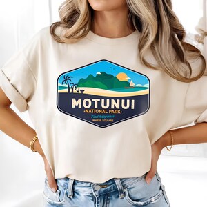 May include: A cream-coloured t-shirt with a graphic of a tropical landscape. The design includes palm trees, mountains, a sailboat, and a setting sun, with the text "MOTUNUI - NATIONAL PARK - Find happiness where you are."