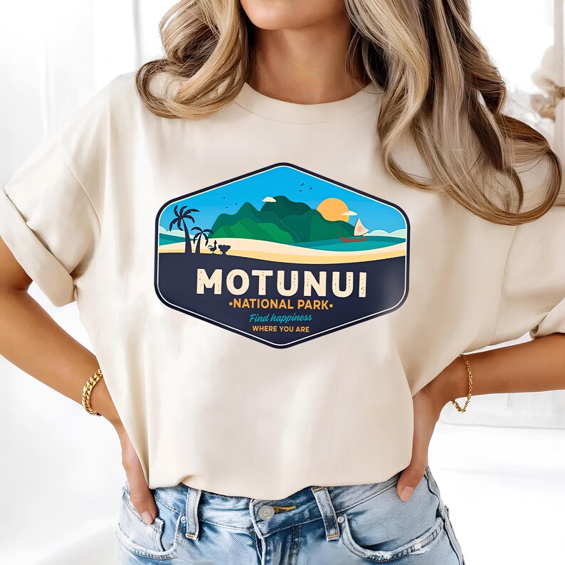 May include: A cream-coloured t-shirt with a graphic of a tropical landscape. The design includes palm trees, mountains, a sailboat, and a setting sun, with the text "MOTUNUI - NATIONAL PARK - Find happiness where you are."