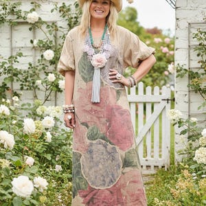 May include: A beige maxi dress with a large floral print in shades of red, pink, and green. The dress has short sleeves and is accessorized with a hat, necklace, and bracelets. The background features a white fence and climbing roses.