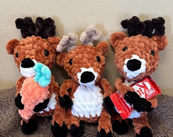 May include: Three brown crocheted reindeer plush toys. The reindeer on the left is holding a carrot, the reindeer in the middle is holding a white and brown plush carrot, and the reindeer on the right is holding a red and white chocolate bar.