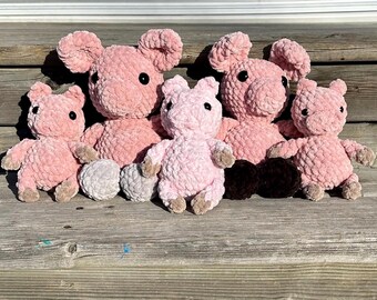 May include: Five pink crocheted pig plushies with black eyes. The plushies are sitting on a wooden surface.