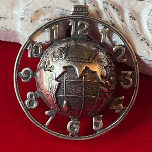 May include: A circular silver-toned pendant featuring a three-dimensional globe at the center. The outer ring of the pendant is designed as a clock face with raised numerals from one to twelve positioned around the central globe sphere.