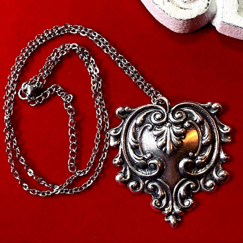 May include: Silver-toned pendant necklace with an ornate, heart-shaped pendant. The pendant features intricate scrollwork and a central floral design. The chain is a delicate, linked style. The necklace is displayed against a vibrant red background.