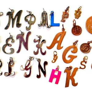 May include: A collection of silver and copper letter charms, including the letters A, C, E, G, H, J, K, L, M, N, P, R, S, T, and Y. The charms are in various styles and sizes.