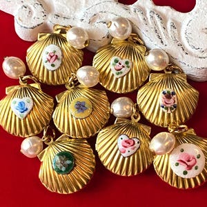 以下が含まれることがあります： 金色の貝殻ペンダントのコレクション。各ペンダントには、さまざまな色の小さな手描きの花柄またはハートのデザインが施されています。ペンダントは赤い表面に配置されています。
