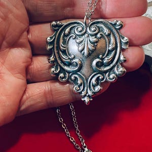 May include: Silver-toned pendant necklace with an ornate, heart-shaped design. The pendant features intricate scrollwork and a central floral motif. The necklace has a delicate chain and a lobster clasp. The pendant is approximately 2 inches in length.