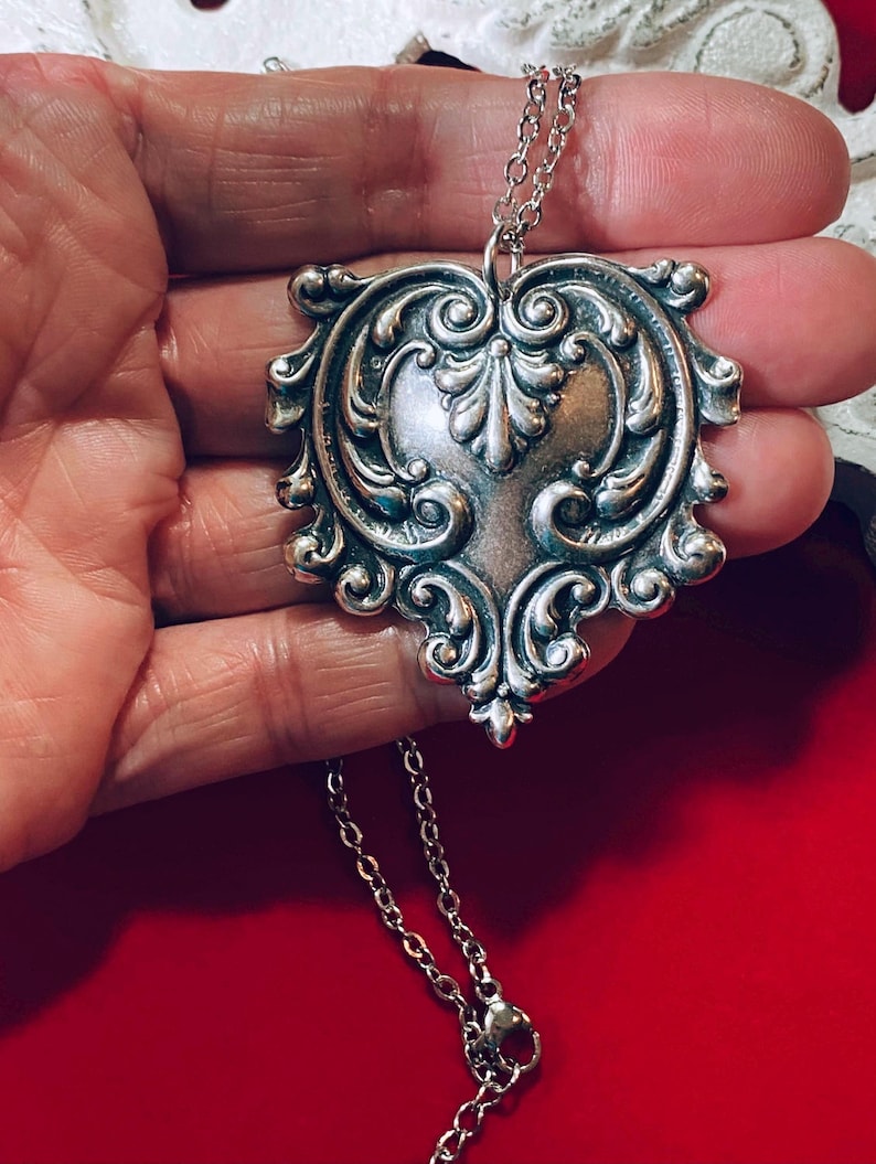 May include: Silver-toned pendant necklace with an ornate, heart-shaped design. The pendant features intricate scrollwork and a central floral motif. The necklace has a delicate chain and a lobster clasp. The pendant is approximately 2 inches in length.