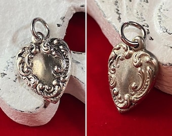 May include: A silver heart-shaped charm with an ornate design. The charm has a loop at the top for attaching to a necklace or bracelet.