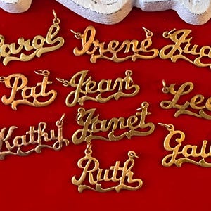 May include: Gold-colored name pendants in various cursive fonts, including "Carol", "Irene", "Judy", "Pat", "Jean", "Lee", "Kathy", "Janet", "Gail", and "Ruth". Each pendant has a small loop for a chain, set against a red background.