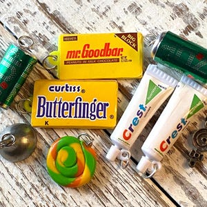 May include: A selection of miniature charms, including a yellow Mr. Goodbar candy bar, a Butterfinger candy bar, two Crest toothpaste tubes, a green soda can, a lollipop, and a silver ball. The charms are on a distressed wooden surface.