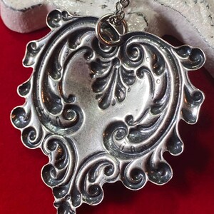 May include: Silver-toned pendant necklace with an ornate, symmetrical design. The pendant features a heart-like shape with detailed scrollwork and floral motifs. The necklace hangs on a delicate chain, creating an elegant accessory.
