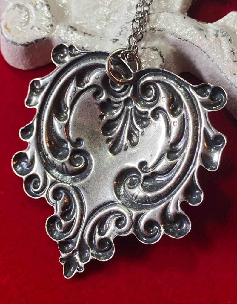 May include: Silver-toned pendant necklace with an ornate, symmetrical design. The pendant features a heart-like shape with detailed scrollwork and floral motifs. The necklace hangs on a delicate chain, creating an elegant accessory.