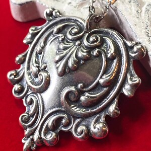 May include: A silver-toned pendant necklace with an ornate, heart-shaped design. The pendant features intricate scrollwork and a central, smooth area. The necklace is displayed against a red background.