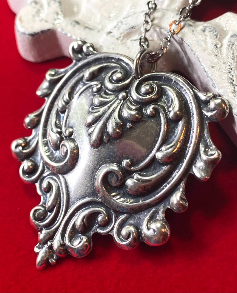 May include: A silver-toned pendant necklace with an ornate, heart-shaped design. The pendant features intricate scrollwork and a central, smooth area. The necklace is displayed against a red background.