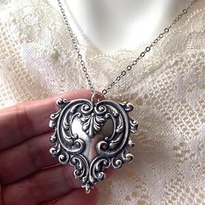 May include: Silver-toned heart-shaped pendant necklace with intricate scrollwork and a delicate chain. The pendant features a detailed, ornate design, perfect for adding a touch of vintage elegance to any outfit.
