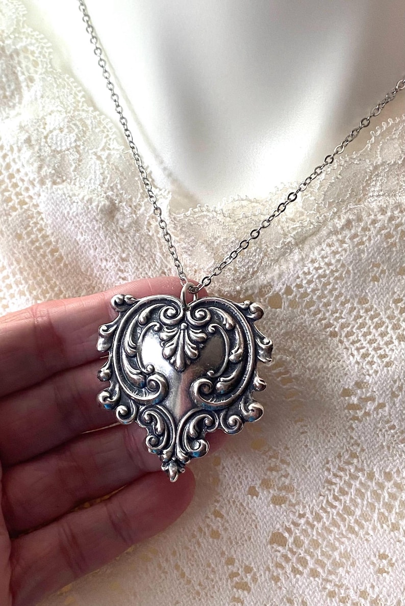 May include: Silver-toned heart-shaped pendant necklace with intricate scrollwork and a delicate chain. The pendant features a detailed, ornate design, perfect for adding a touch of vintage elegance to any outfit.