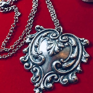 May include: A silver-toned pendant necklace with an ornate, heart-shaped design. The pendant features intricate scrollwork and a central, smooth area. The chain is also silver and visible against a red background.