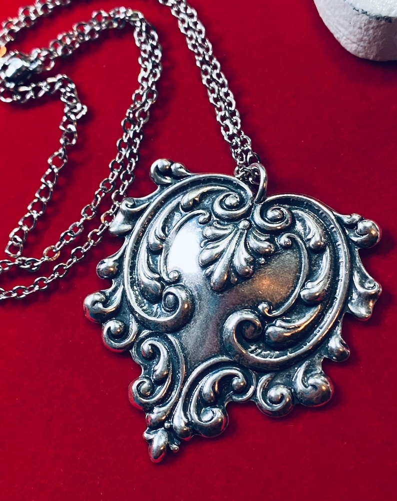 May include: A silver-toned pendant necklace with an ornate, heart-shaped design. The pendant features intricate scrollwork and a central, smooth area. The chain is also silver and visible against a red background.