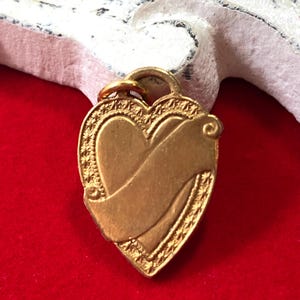May include: A gold-colored heart-shaped charm with a decorative scroll design. The charm has a loop at the top for attaching to a necklace or bracelet.