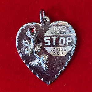 May include: Silver heart-shaped pendant with a rose and the words "I'll Never Stop Loving You." The pendant has a small red gem and a textured edge. The background is a solid red.