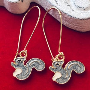May include: A pair of gold-colored drop earrings featuring a gray and white squirrel design. The earrings have a long, curved hook and a detailed squirrel charm. The background is a solid red color.
