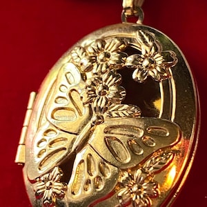 Puede incluir: Colgante de medallón ovalado dorado con diseño de mariposa y flores. El medallón es muy detallado, con una mariposa y flores en relieve. El colgante tiene una abertura con bisagras y un lazo para una cadena.