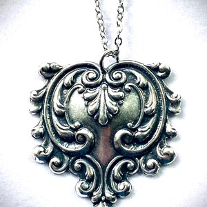May include: Silver-toned pendant necklace with an ornate, symmetrical design. The pendant features a central heart-like shape surrounded by swirling, detailed scrollwork. The necklace chain is also silver-toned and delicate.