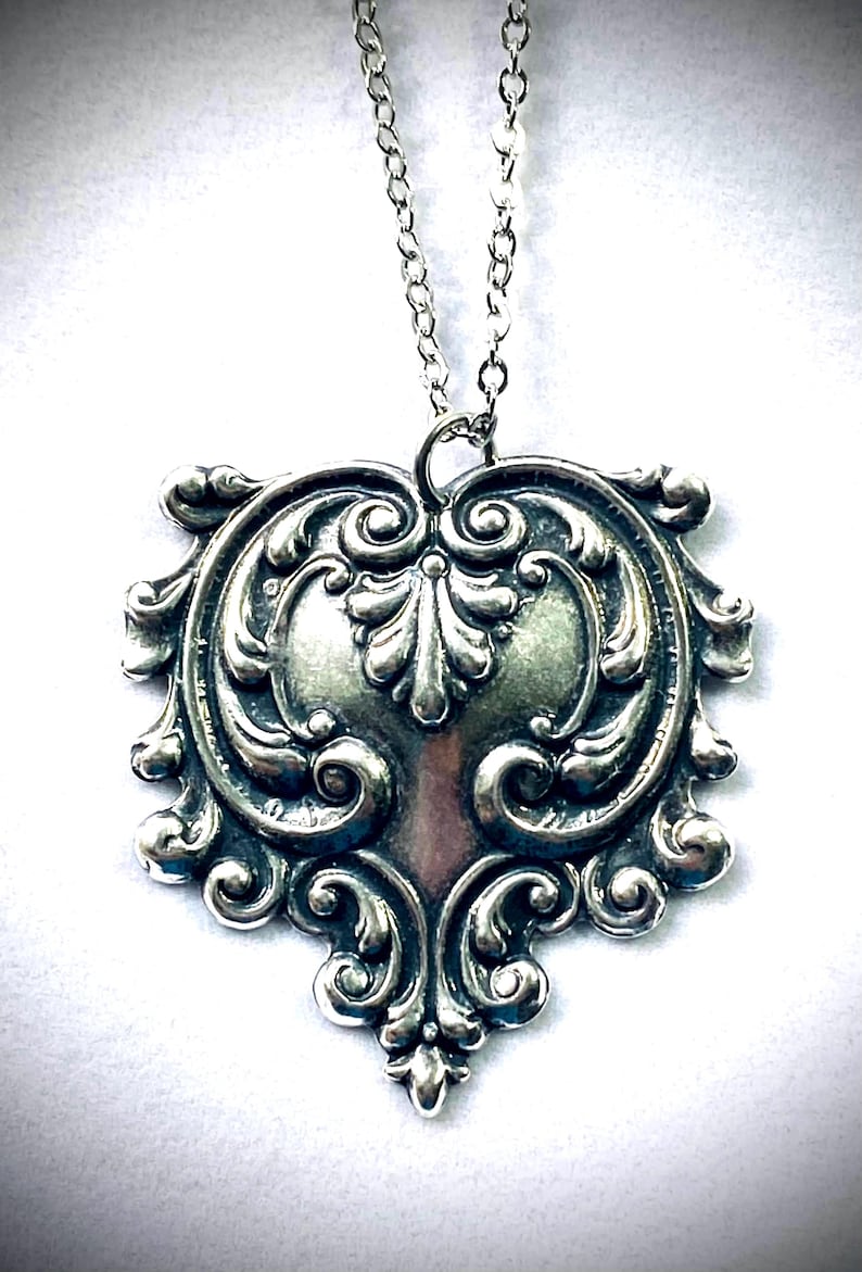May include: Silver-toned pendant necklace with an ornate, symmetrical design. The pendant features a central heart-like shape surrounded by swirling, detailed scrollwork. The necklace chain is also silver-toned and delicate.