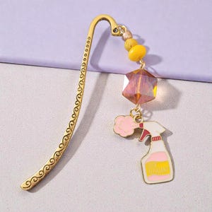 May include: Gold-toned metal bookmark with a decorative swirl design. It features a faceted amber-colored crystal, wooden beads, and a charm of a pink and white spray bottle with the text "DUST REPELLENT".