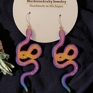 May include: A pair of iridescent snake earrings with gold accents. The earrings are made of acrylic and have a holographic finish. The earrings are handmade in Michigan.