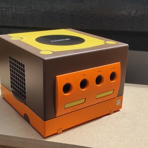 May include: A brown and orange GameCube console with a yellow top and a black disc drive. The console has four controller ports and a memory card slot.