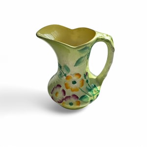 May include: A small, green ceramic jug with a floral design. The jug has a handle and a wide mouth. The flowers are painted in shades of yellow, pink, and purple.