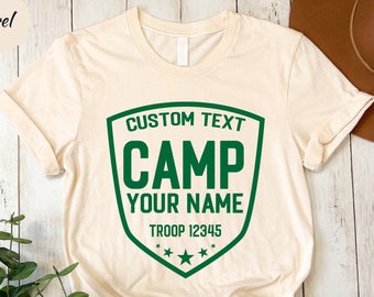 May include: A cream-coloured t-shirt featuring a green shield design. The shield displays the text "CUSTOM TEXT CAMP YOUR NAME TROOP 12345" in green. The t-shirt is knotted at the bottom and has a "very style clothing" logo.