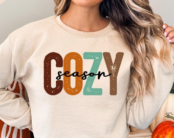 May include: A beige sweatshirt with the text "COZY season" in colorful retro-style letters. The letters are in shades of brown, orange, teal, and brown.