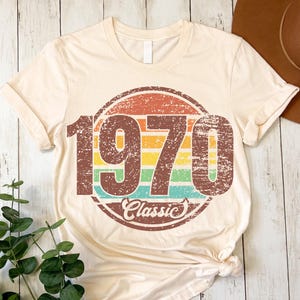 May include: Cream-colored t-shirt with a distressed graphic. The graphic features the number "1970" in a brown, vintage font, set within a colorful circle with the word "Classic" below. The shirt is knotted at the bottom.