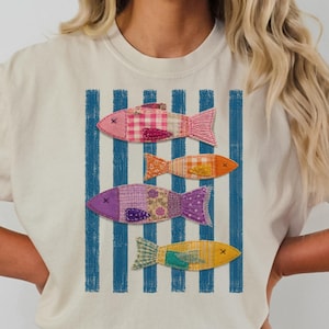 May include: Ivory-colored t-shirt featuring a design of four colorful fish. The fish are in shades of pink, orange, purple, and yellow, with patchwork patterns. Blue vertical stripes form the background.