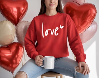 May include: A red sweatshirt with the word "love" in white script and a small heart. The sweatshirt is paired with blue jeans. Heart-shaped balloons in red, pink, and cream are in the background. A white mug is held in the hands.