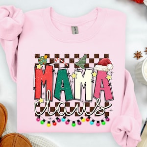 May include: Pale pink sweatshirt with a Christmas-themed "Mama Claus" design. The graphic displays "MAMA Claus" in festive colours, adorned with Christmas decorations, a candy cane, a Christmas tree, and a Santa hat. A string of colourful lights is at the bottom.