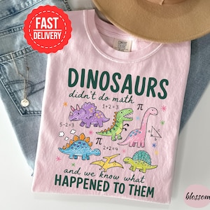 A pink t-shirt with a cartoon dinosaur design and text, along with a hat and jeans, is displayed on a table.