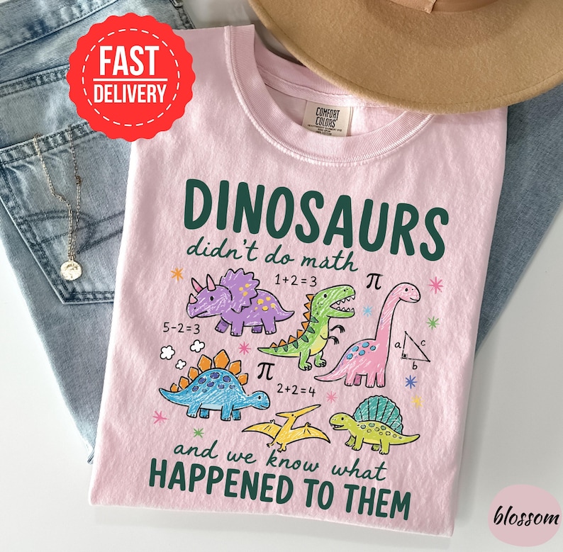 A pink t-shirt with a cartoon dinosaur design and text, along with a hat and jeans, is displayed on a table.