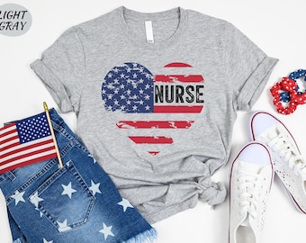 Puede incluir: Una camiseta gris claro con un diseño de corazón de bandera estadounidense desgastado que dice "NURSE" en texto negro.